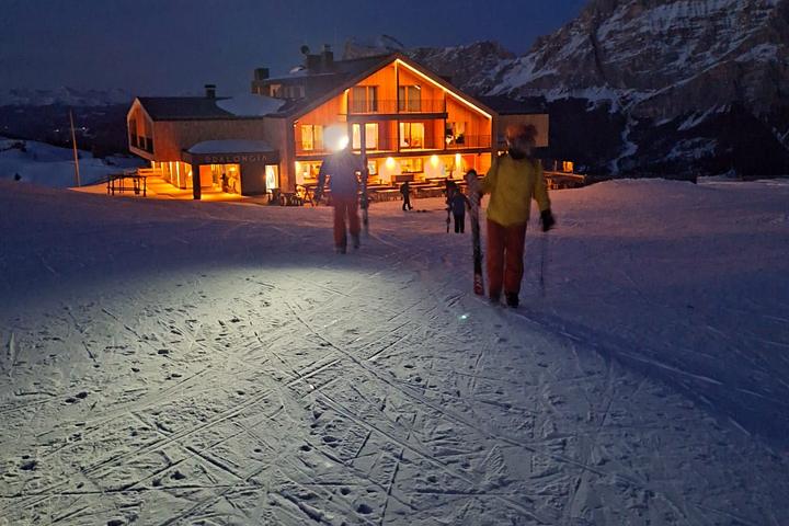 Skiers walking to a lit mountain hut at dusk in the Dolomites